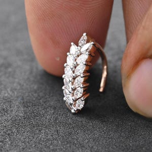 May include: A rose gold nose ring with a cluster of marquise-cut white gemstones. The gemstones are arranged in a leaf-like pattern, creating a delicate design. The jewellery is held between two fingers against a dark grey background.