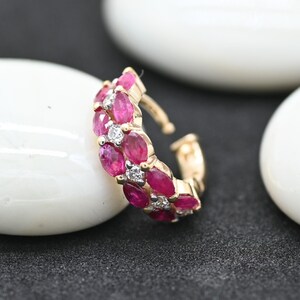 May include: A gold-toned ear cuff featuring a double row of oval-cut, vibrant pink gemstones, interspersed with small, round, clear gemstones. The jewelry piece is set against a dark background with white, rounded objects.