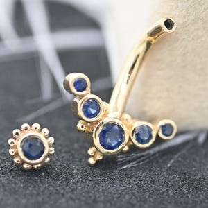 Op de afbeelding: Goudkleurige sieradenset met een gebogen barbell en een kleine stud, beide versierd met levendige blauwe edelstenen. De barbell heeft meerdere edelstenen, terwijl de stud een enkele, centrale steen heeft omringd door kleinere accenten.