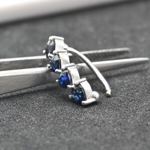 Op de afbeelding: Een zilverkleurige oormanchet met drie donkerblauwe edelstenen. Het gebogen ontwerp heeft kleine, rechthoekige zettingen die de stenen vasthouden. De oormanchet wordt getoond tegen een donkere achtergrond met een zilveren pincet.