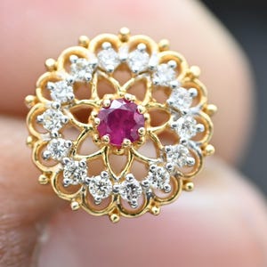 May include: A gold-toned floral-shaped jewelry piece with a central ruby gemstone and surrounding diamonds. The design features a detailed filigree pattern, creating a delicate and elegant appearance. This piece is likely a stud earring or a similar small jewelry item.