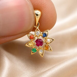 May include: A gold pendant with a flower design, featuring a central ruby surrounded by colorful gemstones. The pendant is held up by a finger against a satin background. The gemstones include a diamond, blue sapphire, pearl, orange sapphire, green emerald, and a moonstone.