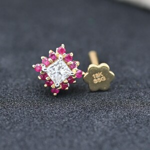May include: A gold stud earring featuring a square, clear gemstone at its centre, encircled by small, red gemstones. The earring has a flower-shaped backing, marked with "18K SSG". The earring is set against a dark, textured backdrop.