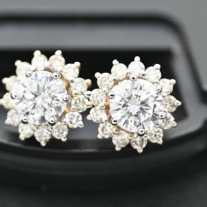 Puede incluir: Un par de pendientes de diamantes. Cada pendiente presenta un diamante grande, redondo y transparente rodeado de diamantes más pequeños, creando un halo en forma de flor. Los pendientes están engarzados en metal dorado.