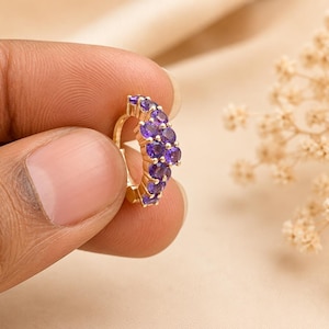 May include: A gold-colored hoop earring with a row of small, round, purple gemstones. The earring is held between fingers against a soft, neutral background. The gemstones are a deep, rich purple color.