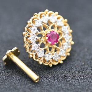Pode incluir: Um piercing de nariz dourado com um design floral. O piercing apresenta uma gema de rubi central rodeada por pequenas gemas transparentes. A haste é dourada e tem uma pequena extremidade em forma de flor.