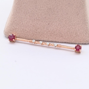 May include: A rose gold industrial barbell piercing jewelry featuring a round red ruby gemstone at each end and a central section decorated with four small, clear, round-cut white diamonds set in a delicate, twisted metal bar design.