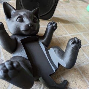 May include: A black cat-shaped figurine, lying on its back with its paws raised. The figurine has a gray stripe down its back and is made of a textured material. The cat is a decorative item.