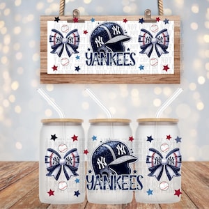 May include: A wooden sign and three glass tumblers featuring a New York Yankees theme. The sign displays a baseball helmet, bow, and the word "Yankees." The tumblers have the same design, with a bamboo lid and a straw.