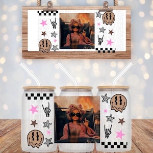 May include: A glass tumbler with a bamboo lid and clear straw featuring a graphic print of a doll in a pink robe and sunglasses, surrounded by black and white checkered patterns, pink stars, brown stars, skeleton hand signs, and melting smiley faces.
