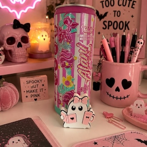 May include: A tall Alani Nu energy drink can with a pink and white striped label featuring graphics of a balloon dog, cotton candy, and a rubber duck. In front of the can sits a small white ghost figurine holding a pink book and a pink cup.