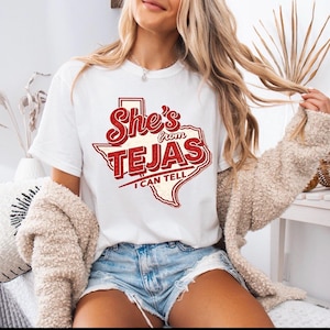 May include: White t-shirt with a red and white graphic that reads "She's from Tejas I can tell" in a stylized font. The graphic is in the shape of the state of Texas. The shirt is a crew neck and has short sleeves.