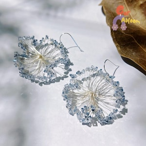 May include: A pair of handmade earrings with a floral design. The earrings have a silver wire frame with a central cluster and radiating wires, decorated with light blue crystals around the edges. The earrings are shown on a white surface.