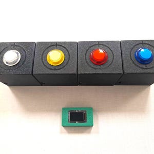 May include: Four black square boxes with clear, yellow, red, and blue buttons. Each box has a circular design around the button. Below is a small green rectangle with a screen and the text "VBS".