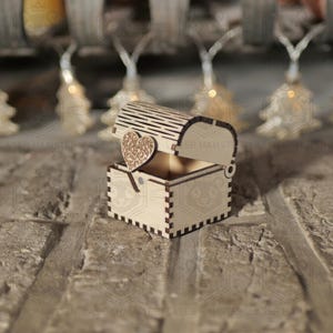 Puede incluir: Un pequeño cofre del tesoro de madera abierto con un adorno en forma de corazón. El cofre está hecho de madera de color claro y presenta un corazón decorativo en la tapa. El cofre está sobre una superficie de ladrillo.