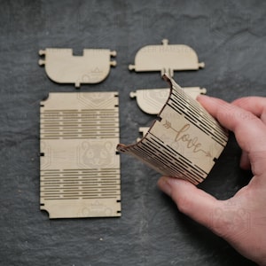 Può includere: Una scatola di legno smontata con dettagli tagliati al laser. I componenti della scatola sono marrone chiaro e includono un lato curvo con la parola "love" e un design a freccia. Le altre parti sono piatte e rettangolari.