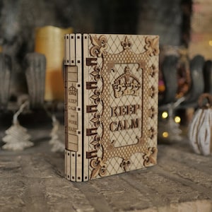 May include: A wooden box with a book design. The box is light brown with intricate laser-cut details, including a crown and the words "KEEP CALM" and "WORK HARD". The box has a vintage, decorative style.
