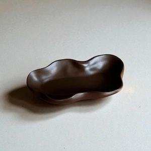 May include: A dark brown ceramic dish with an organic, flowing shape. The dish has a smooth, glossy surface and a shallow, irregular form, suitable for serving small items or as a decorative piece. It is set against a neutral background.