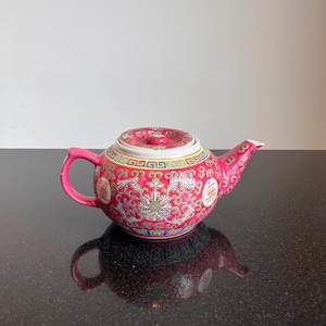 Può includere: Piccola teiera rosa ornata con coperchio abbinato. Presenta un design floreale dettagliato con dettagli dorati e caratteri cinesi. Il manico e il beccuccio sono rosa acceso, su una superficie scura.