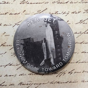 May include: A round, black and white button featuring a vintage image of a rocket on a launch pad. The button has the text "AEROJET" at the top and "BEYOND TODAY TOWARD TOMORROW" around the edge.