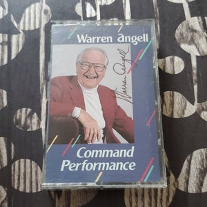 May include: A cassette tape with a photo of Warren Angell, a man with glasses, smiling and wearing a red jacket. The text "Warren Angell" is at the top of the cassette tape and "Command Performance" is at the bottom.