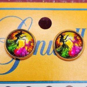 May include: Two round gold-toned earrings with a colorful witch design. The earrings feature a silhouette of a witch in a black hat and dress, with a vibrant background of yellow, green, pink, and orange. The earrings are set against a yellow background.