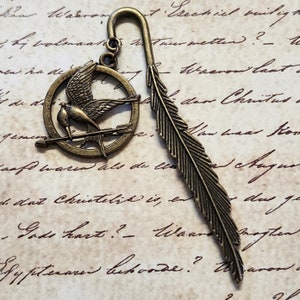 May include: A bronze bookmark featuring a stylized mockingjay bird in a circle with an arrow, and a feather design on the other side.