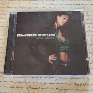 May include: CD case for Alicia Keys' album "songs in A minor." The cover features a woman in a black leather jacket and a green hat. The album title and artist's name are in white text.