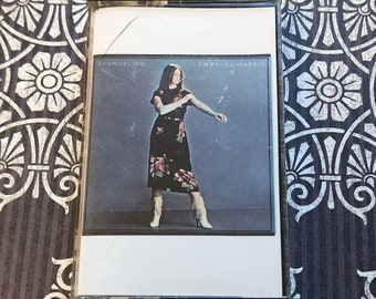 May include: A cassette tape case with a photo of a woman wearing a black and white dress and white boots. The text "Evangeline" and "Emmylou Harris" is printed on the case.