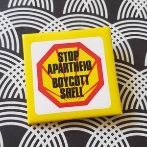 May include: A yellow square button with a red stop sign featuring a white Shell logo and the text "STOP APARTHEID BOYCOTT SHELL".