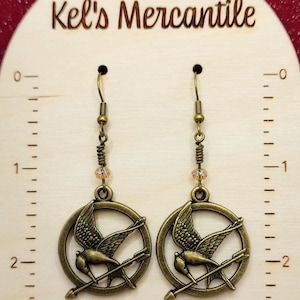 May include: A pair of bronze earrings with a mockingjay design. Each earring features a circular pendant with a stylized mockingjay bird and an arrow, suspended from a bronze wire with a small crystal bead.