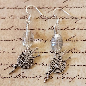 May include: Silver-toned dangle earrings featuring a clear, oval-shaped bead and a charm depicting a ball of yarn with knitting needles. The earrings have a vintage aesthetic, perfect for a crafter or knitter.