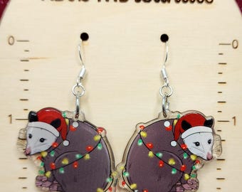May include: Dangle earrings with opossum designs wearing Santa hats and Christmas lights. The earrings are made of clear acrylic with silver-coloured hooks. The background is a wooden surface with the text "Kel's Mercantile".