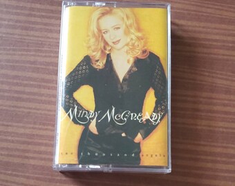 May include: A Mindy McCready cassette tape with a yellow background. The cover features a photo of a woman with blonde hair wearing a black top. The text reads "Mindy McCready" and "ten thousand angels".
