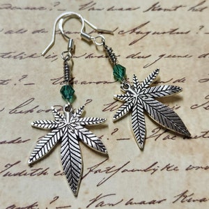 May include: Silver marijuana leaf earrings with green crystal accents. The earrings are hanging from silver hooks.