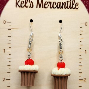 May include: A pair of dangle earrings featuring miniature chocolate cupcakes with white frosting and red cherries. The earrings are silver with clear and amber beads.