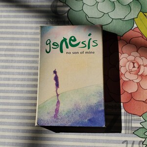 May include: A vintage cassette tape case with a watercolor-style image of a person standing on a hill. The case is for the Genesis album "No Son of Mine".