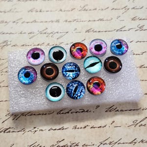May include: Assortment of 12 colorful glass eyes with various designs, including blue, red, pink, brown, and black. The eyes are arranged on a white foam surface.