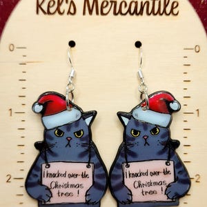 May include: A pair of dangle earrings featuring a cartoon cat wearing a Santa hat and holding a sign that says "I knocked over the Christmas tree!" The cat is blue and gray with a white face and black eyes. The earrings are silver.