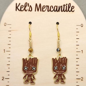 May include: A pair of gold dangle earrings featuring a brown and gold cartoon character with a brown head and a gold body. The character has a brown face with two white eyes and a small mouth. The earrings are hanging from a gold hook with a small gold bead and a black bead.