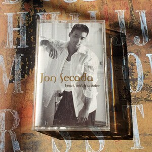 May include: A cassette tape case with a photo of Jon Secada on the front. The text "Jon Secada" is in gold lettering and the text "heart, soul & a voice" is in smaller white lettering.