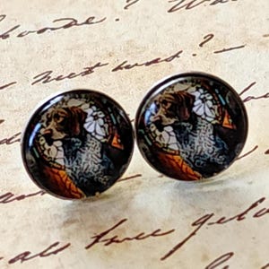 May include: A pair of round stud earrings featuring a detailed illustration of a dog. The earrings have a dark background with brown, white, and orange accents. The earrings are set against a vintage paper background.