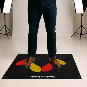 Op de afbeelding: Zwarte rechthoekige PosePoint Pro Mat met abstracte vormen in rood, geel en blauw. Een persoon in een spijkerbroek en bruine schoenen staat op de mat. De tekst "PosePoint Pro Mat" is zichtbaar.