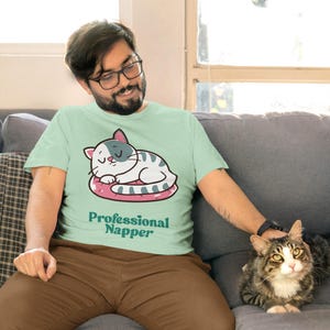 May include: A light green t-shirt with a cartoon cat design and the words "Professional Napper". The cat is white and gray, sleeping on a pink cushion. The person is wearing brown pants.