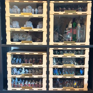 Op de afbeelding: Een modulair houten opbergsysteem voor miniatuurfiguurtjes, bestaande uit meerdere gestapelde lades met doorzichtige fronten. Elke lade is voorzien van labels met teksten zoals Solar Guard, Living Metal en Night Goblins, waarin diverse beschilderde en onbeschilderde spelmodellen zijn georganiseerd.