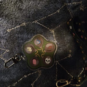 May include: A clear, five-lobed keychain charm with a green background and various small illustrations. The charm has a silver-colored clasp. A black lanyard with a gold-colored clasp is also visible.