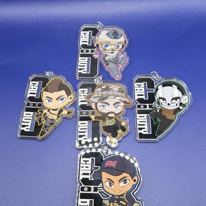May include: Five Call of Duty character keychains. Each keychain features a different character in a cartoon style, with the words "Call of Duty" printed on a black and white background. The characters are wearing military-style outfits and helmets.