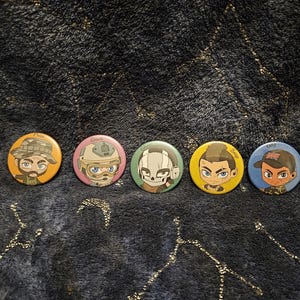 May include: Five round, cartoon-style pinback buttons with character illustrations. The buttons feature different characters with various hairstyles and headwear, including a military hat, helmet, and baseball cap. Each button has a unique color scheme.