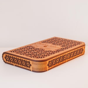 Puede incluir: Una caja rectangular de madera con un diseño geométrico cortado con láser. La caja es de color marrón claro con un patrón floral decorativo en la tapa y los lados. La tapa está grabada con los nombres "Anna John" y la fecha "12 de dic. de 2025".