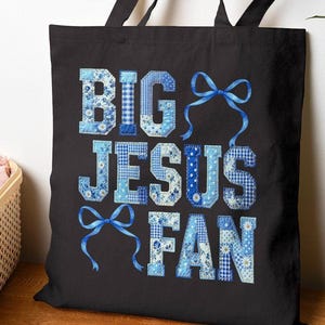 May include: Black tote bag with the text "BIG JESUS FAN" in blue patchwork letters and blue ribbon accents. The bag has black handles and is displayed on a wooden surface.
